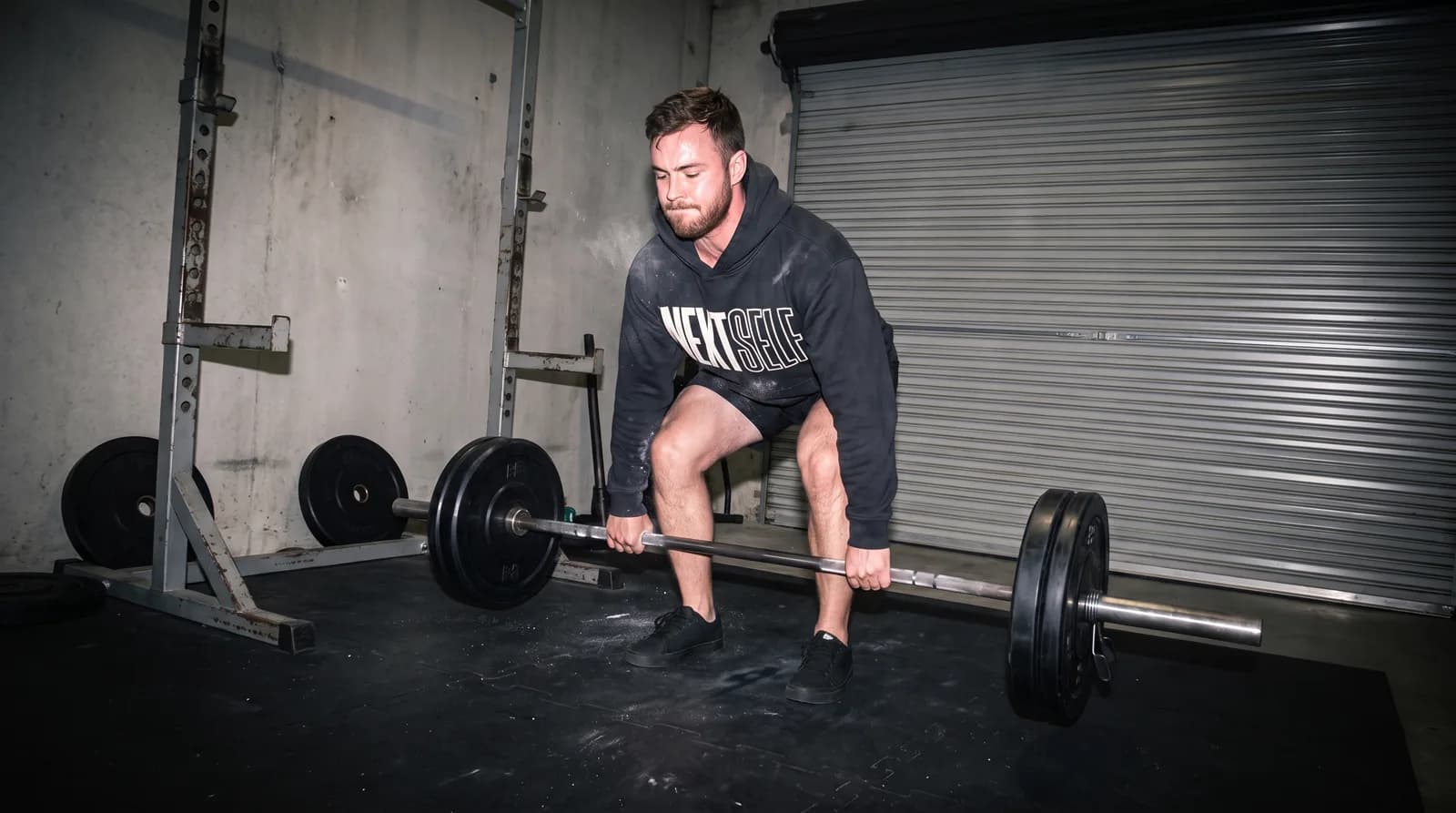 A lifter at deadlift lockout in a garage gym at night, wearing the black Outline Hoodie